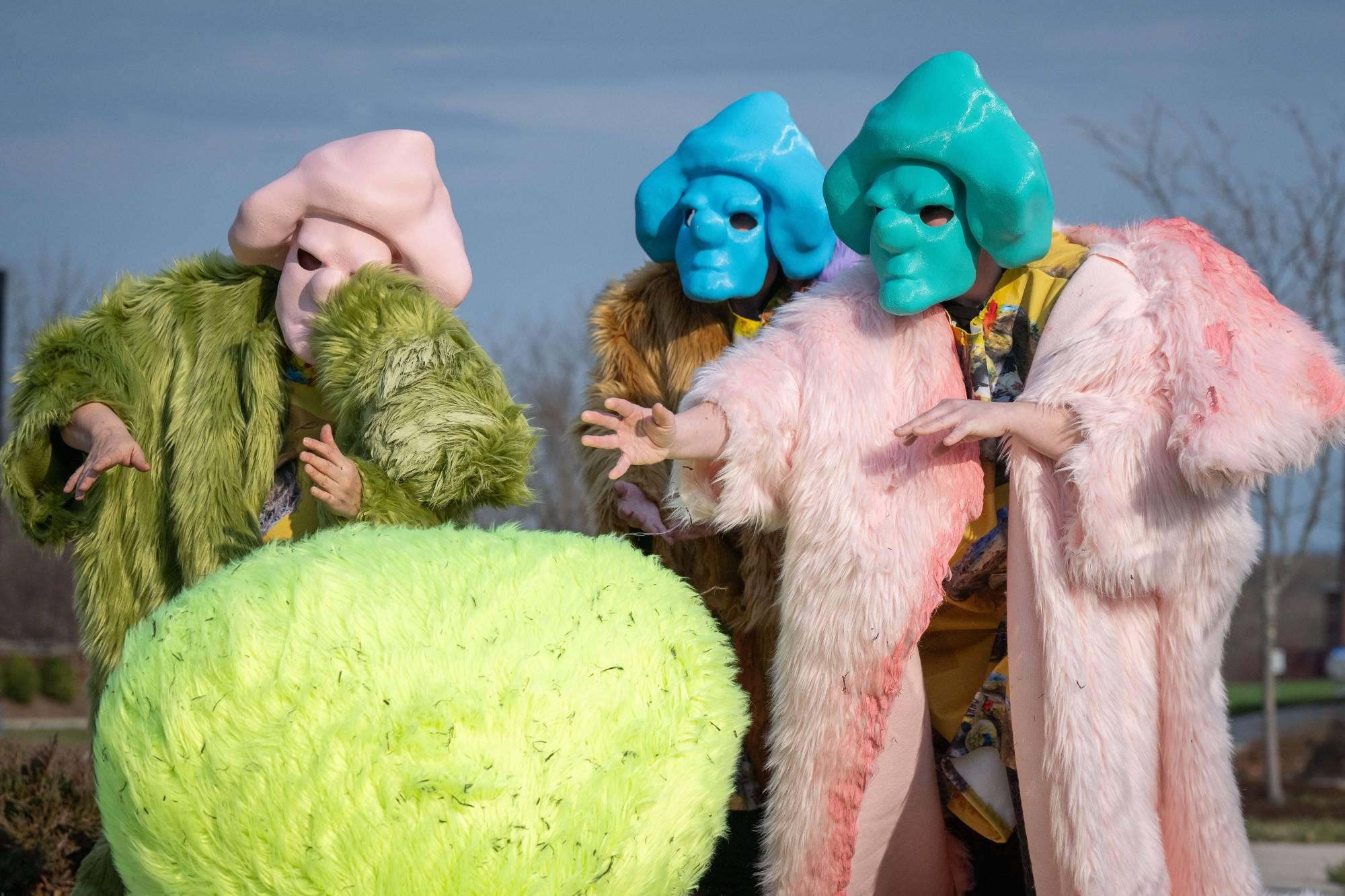 Yellow furry orb with three eerie figures wearing pink blue and green masks.